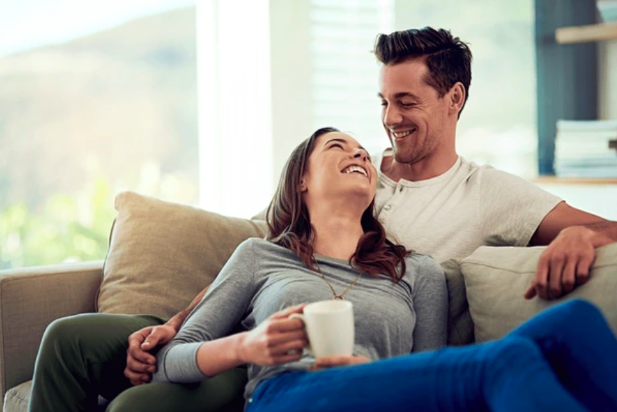 A couple sitting on a couch, laughing and talking while sharing a coffee, showcasing warmth and connection.