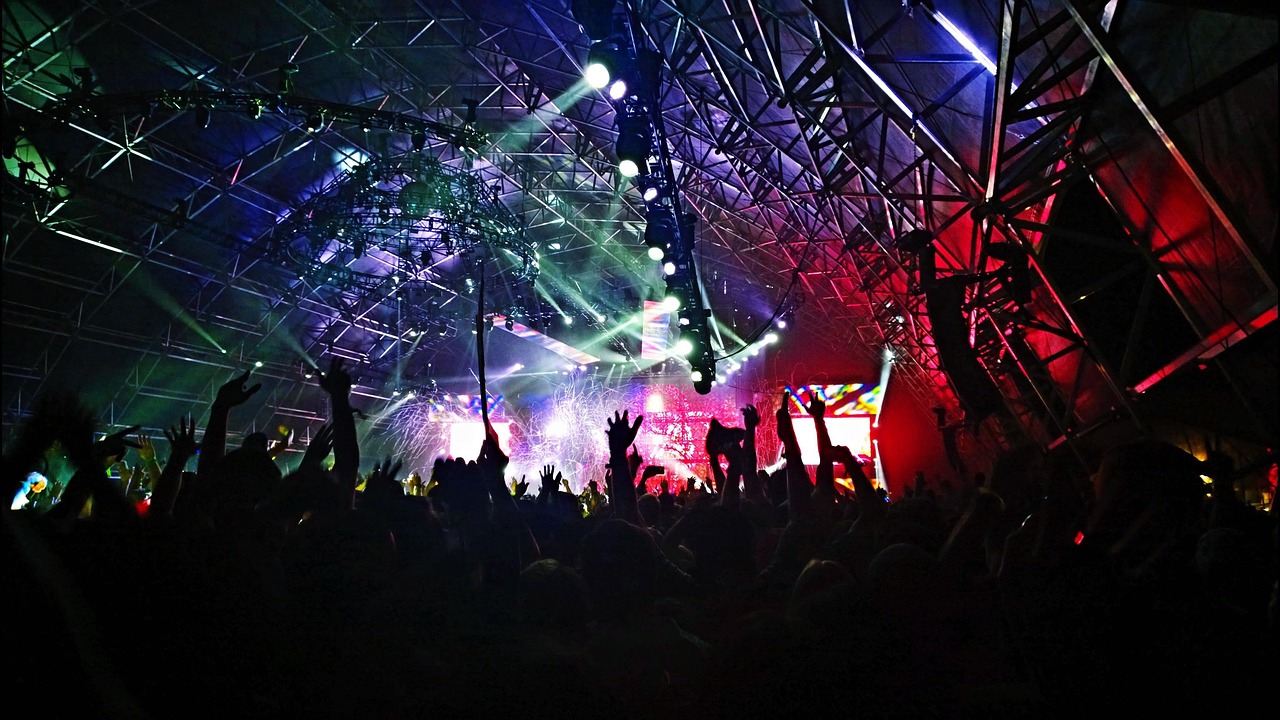 Large crowd raising hands at an indoor music concert with colorful stage lighting and laser effects