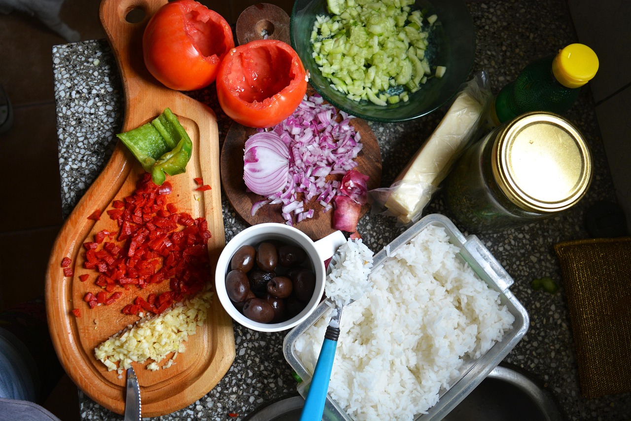 A top view of fresh chopped vegetables, rice, olives, and seasonings arranged on a kitchen counter for cooking.
